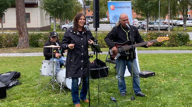 en kvinna sjunger och två män spelar trummor respektive bas utomhus.