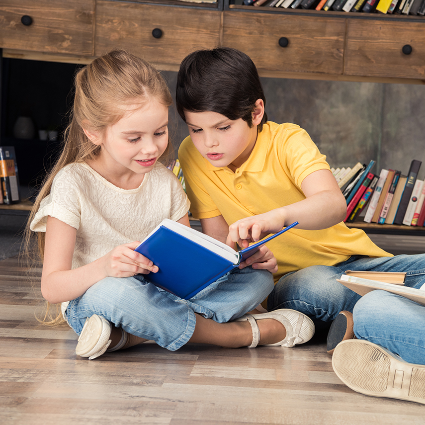 Två barn, en pojke och en flicka sitter på golvet i ett bibliotek och läser böcker
